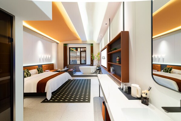 A modern hotel room with a large bed, a separate sitting area, a bathtub, and a vanity with a mirror.