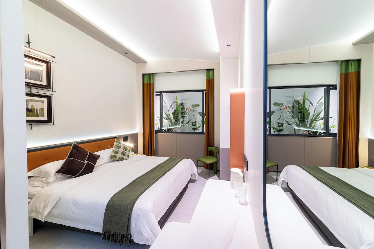 Une chambre d’hôtel avec deux lits, une grande fenêtre avec des rideaux, un fauteuil vert et une table de chevet avec une lampe.