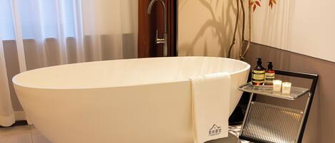 A modern bathroom with a large oval bathtub, a wooden door, a wall-mounted shelf with toiletries, and a potted plant.