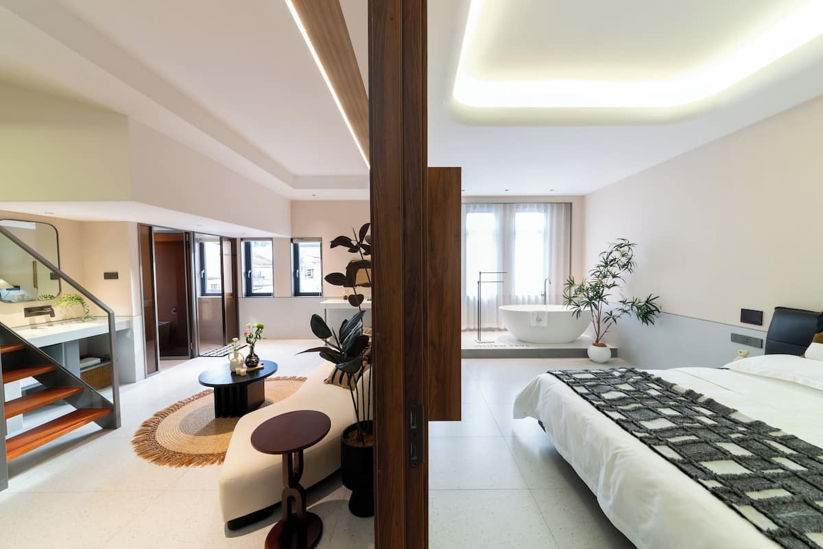 Une chambre d’hôtel moderne comprenant un lit, un coin salon avec une table basse et des fauteuils, une grande fenêtre et une salle de bain avec baignoire.