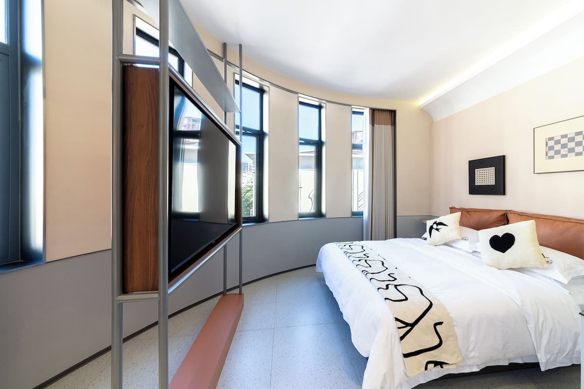 Une chambre d’hôtel moderne avec un lit aux lignes courbes, de grandes fenêtres, un téléviseur et une œuvre d’art à motifs à carreaux accrochée au mur.