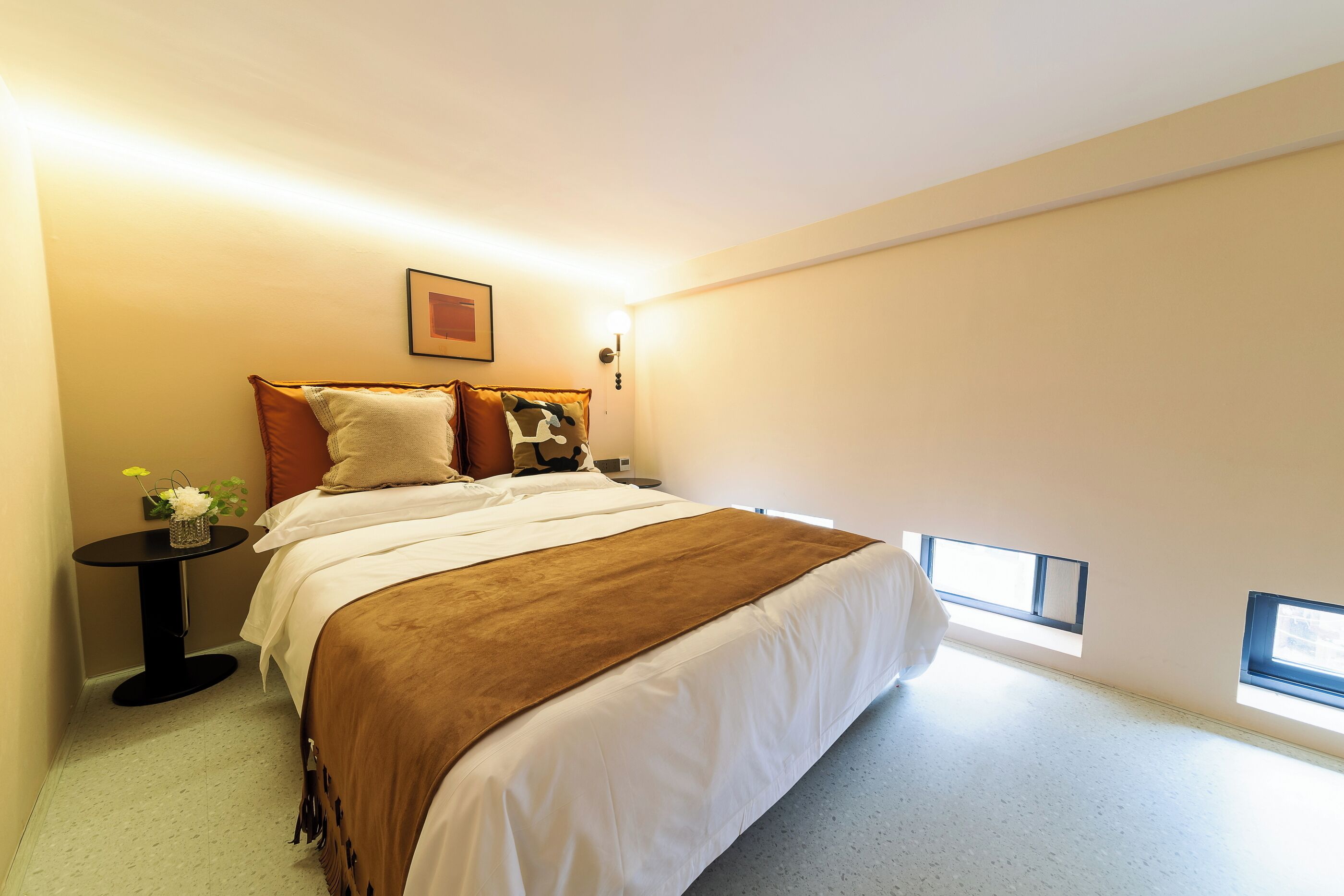 Un dormitorio con cama, mesita de noche, una pequeña mesa con flores y dos ventanas.