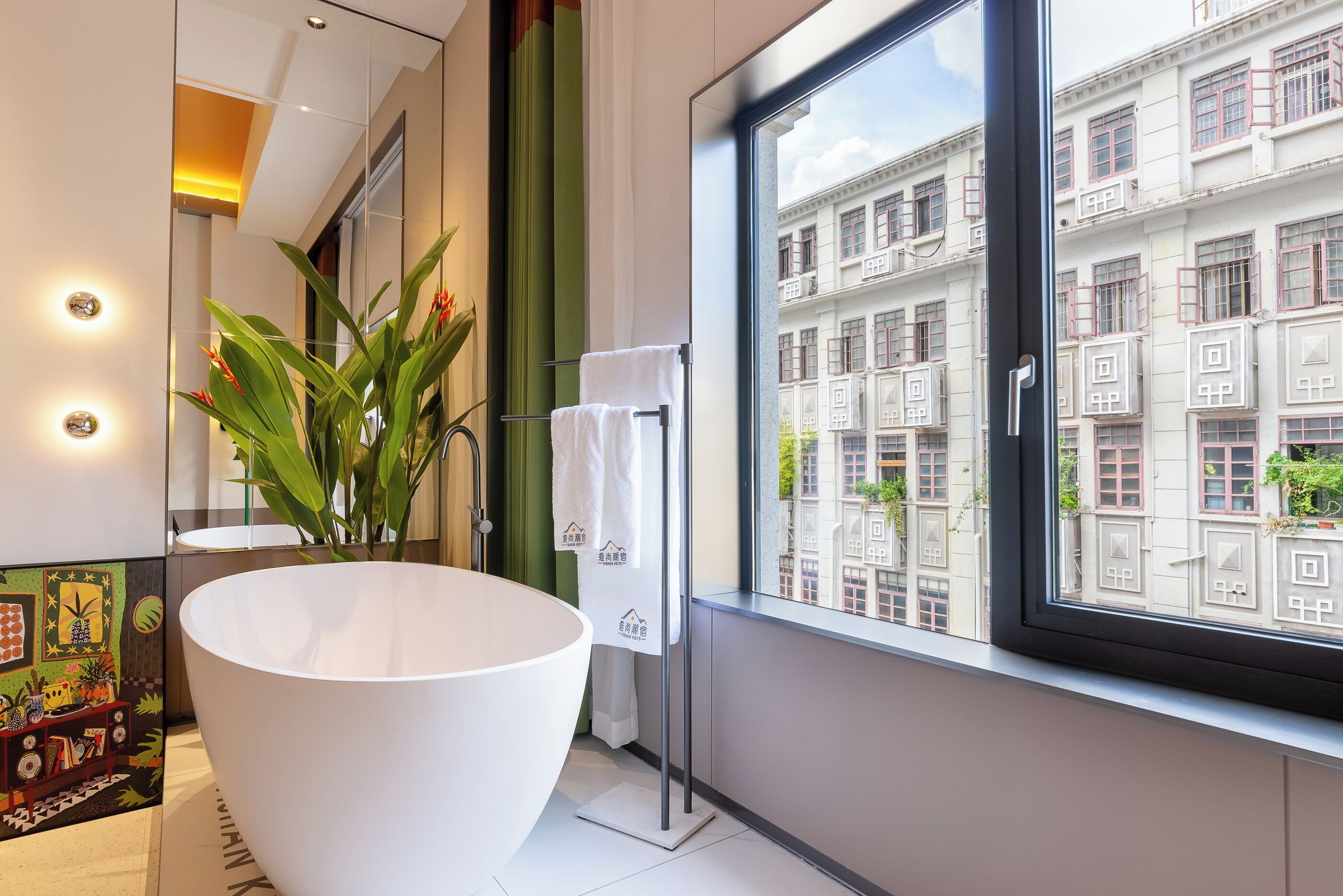 A modern bathroom with a large white bathtub, a potted plant, and a view of a cityscape through a large window.