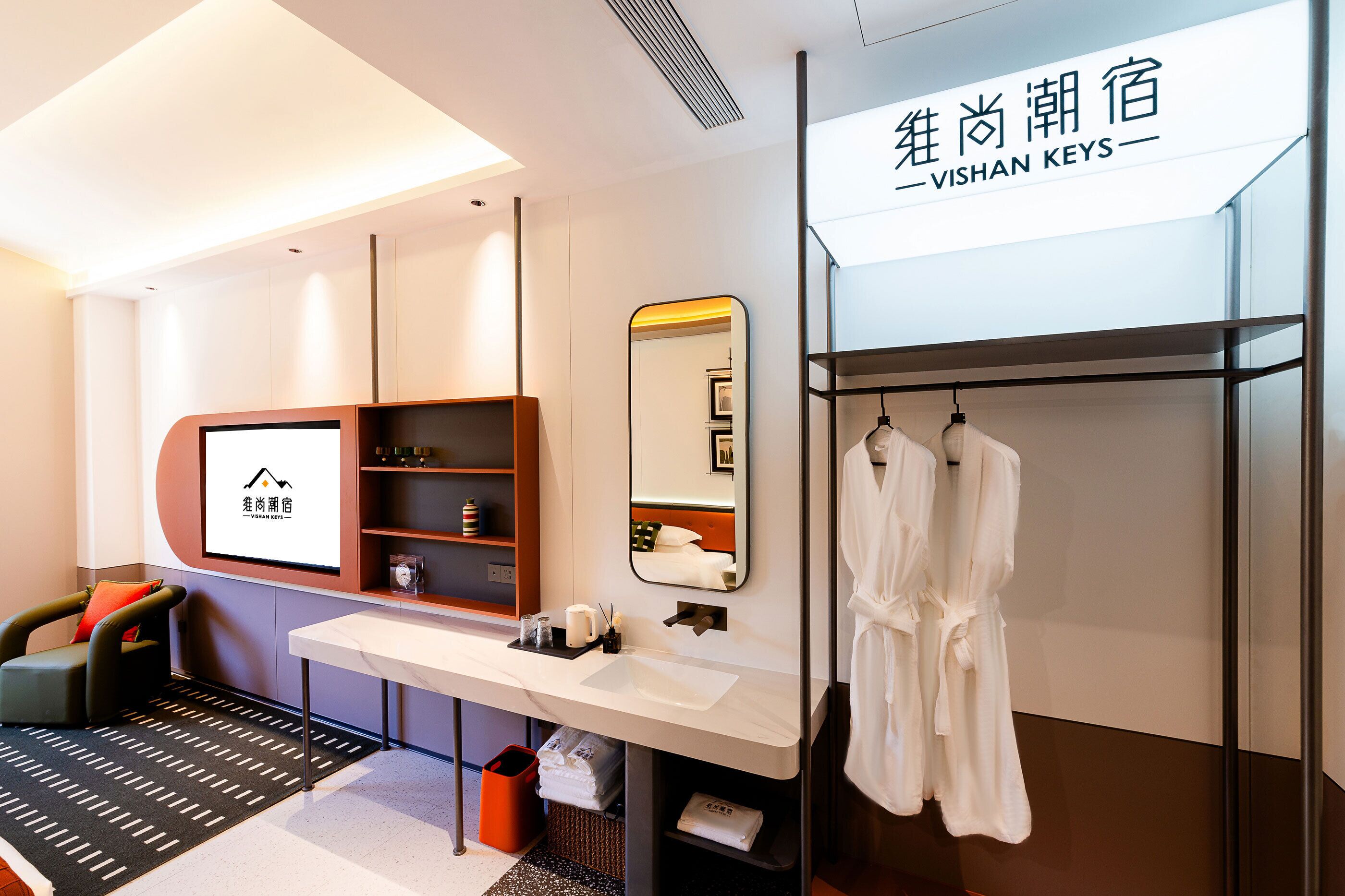 Ein modernes Hotelzimmer mit einem weißen Schminktisch, einem großen Spiegel, einem Bademantel an einem Gestell, einem Massagesessel und einem Fernseher, der den Namen des Hotels anzeigt.