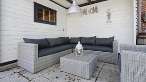 A covered outdoor seating area with a sectional sofa and a square coffee table, featuring a lantern and a wall-mounted fish decoration. - Sunshine Bungalow in Noordwijkerhout (Noordwijkerhout)