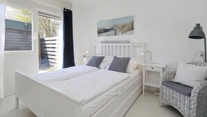 A bedroom with a white bed, a nightstand, a wicker chair, a painting on the wall, and a window with a view of trees. - Sunshine Bungalow in Noordwijkerhout (Noordwijkerhout)