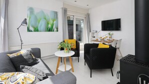 A modern living room with a sofa, armchairs, a small table with a vase of flowers, a TV mounted on the wall, and a black stove. - Sunshine Bungalow in Noordwijkerhout (Noordwijkerhout)