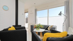 A modern living room with black leather sofas, a round white table with a vase of flowers, a black heater, and a large window with curtains. - Sunshine Bungalow in Noordwijkerhout (Noordwijkerhout)