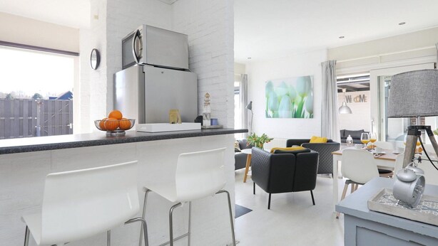A modern kitchen with a large island, white bar stools, and a dining area with a sofa and chairs. - Sunshine Bungalow in Noordwijkerhout (Noordwijkerhout)