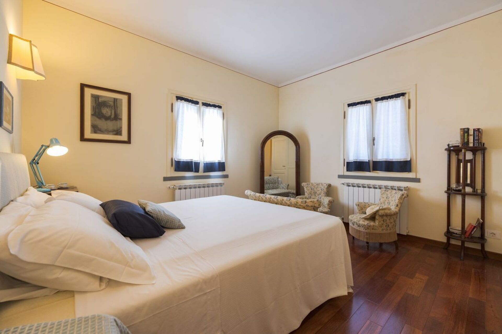 A bedroom with a bed, two chairs, a mirror, and a shelf with books.
