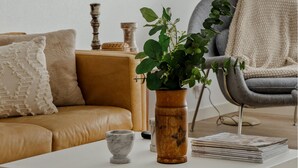 Un salon avec un canapé beige, une table basse ornée d’un vase contenant des plantes vertes, une pile de magazines et un fauteuil gris.