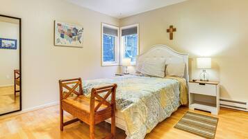 Un dormitorio con una cama, una silla de madera, una mesita de noche con una lámpara, una cruz en la pared y una ventana con persianas.