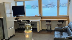 A room with a clock, a TV, a refrigerator, a heater, a couch, and large windows with a view of a snowy landscape. - Train Line Field (Tobetsu)