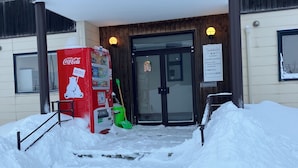 A building entrance with a Coca-Cola vending machine, a green broom, and a snow-covered path leading to the door. - Train Line Field (Tobetsu)