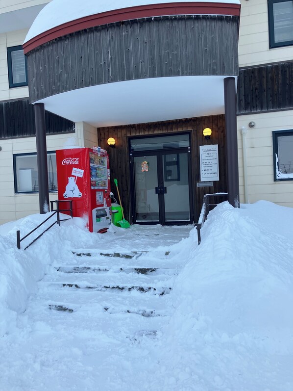 A building entrance with a Coca-Cola vending machine, a green broom, and a snow-covered path leading to the door.