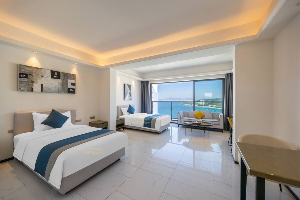 A modern hotel room with two beds, a balcony with a city view, and a dining area.