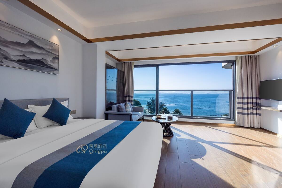 A modern hotel room with a large bed, a sofa, a small table, and a large window offering a view of the sea.