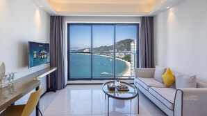 Habitación de hotel moderna con un amplio ventanal con vistas al mar, un sofá con cojines, una mesita con bandeja y un televisor de pantalla plana montado en la pared.