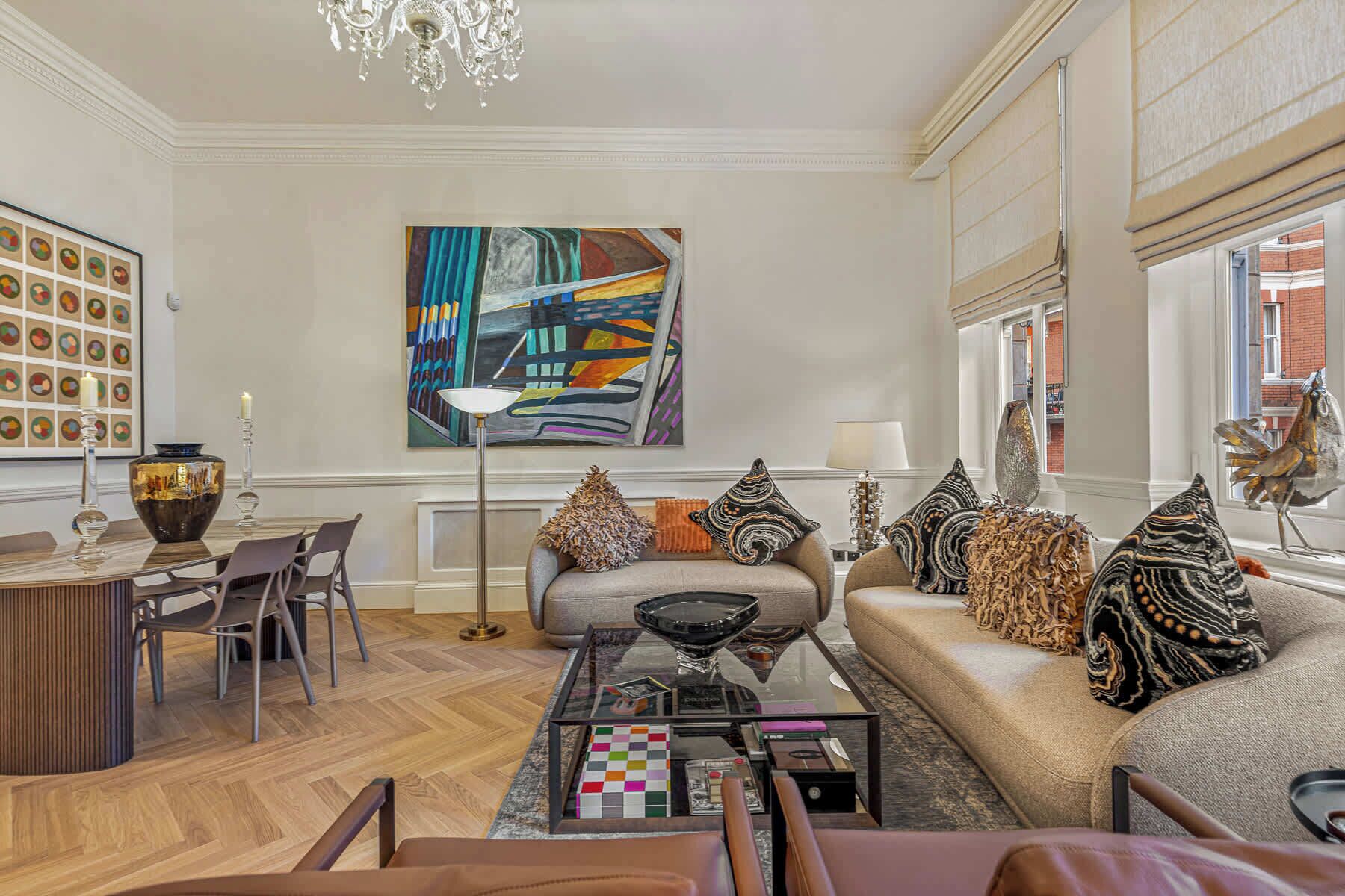 A modern living room with a sofa, coffee table, and chairs. There is a large abstract painting on the wall, a chandelier, and a window with a view of a building.