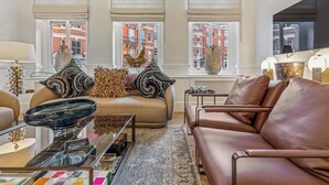A modern living room with a leather sofa, a glass coffee table, and a view of red brick buildings through large windows. - The Gallery - Exquisite Apartment, Kensington (Kensington)