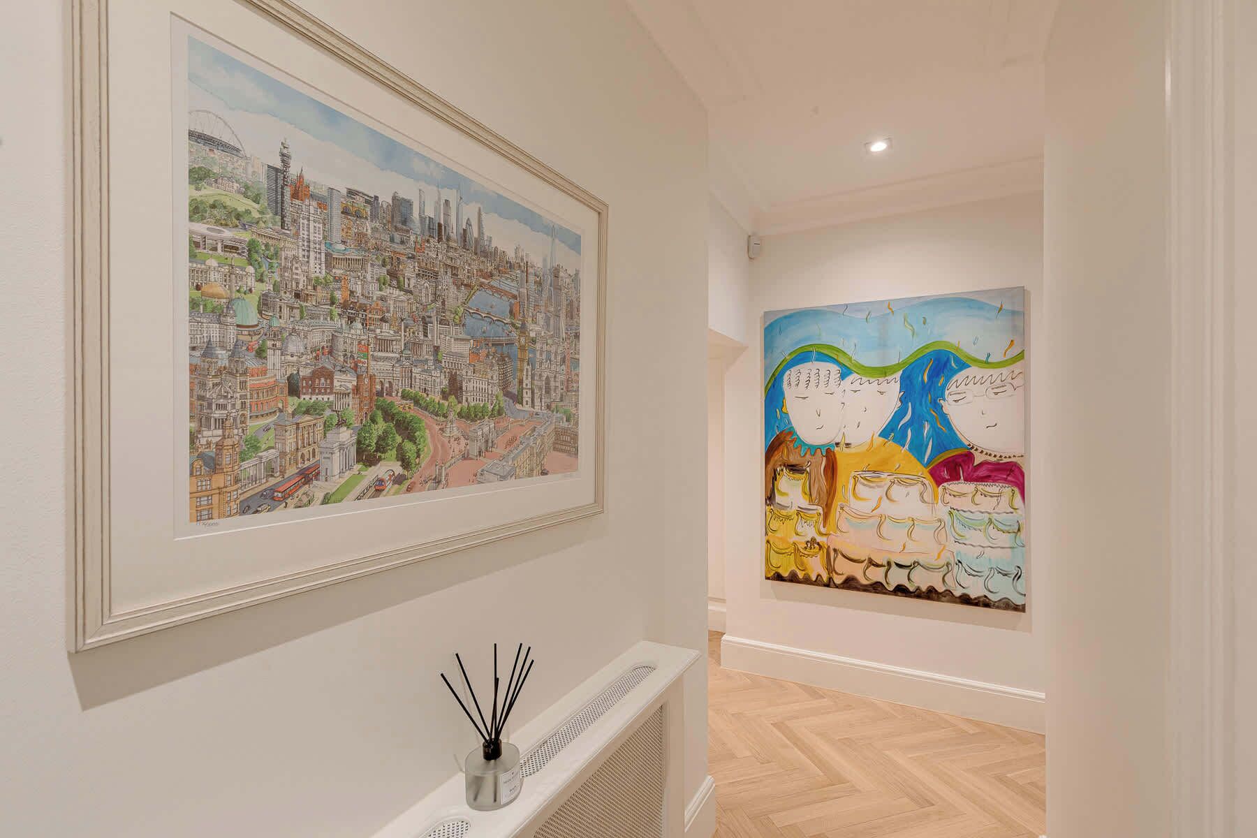 Ett konstgalleri med två inramade målningar på väggen, ett element och en liten vas med dekorativa pinnar.