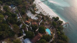 Vista aérea de un complejo turístico costero con playa, piscina y muelle que se adentra en el agua.