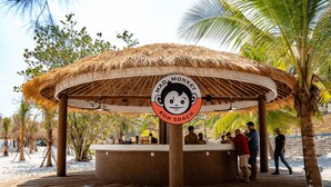 Estructura de techo de paja con una zona de estar circular, rodeada de palmeras y una playa de arena.