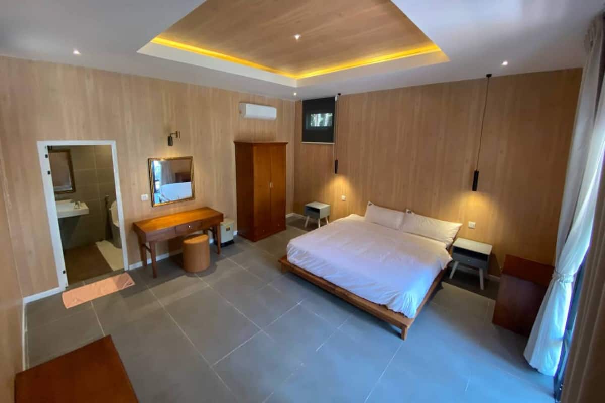A modern bedroom with a wooden ceiling, a large bed, a desk, a wardrobe, and a TV mounted on the wall.