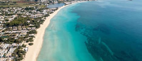 Vista aérea de una zona costera con una extensa playa de arena, aguas turquesas cristalinas y una zona residencial densamente poblada con casas y edificios.