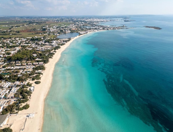An aerial view of a coastal area with a long sandy beach, clear turquoise waters, and a densely populated residential area with houses and buildings.