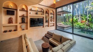 A modern living room with a beige sofa, a TV mounted on the wall, and a large window overlooking a garden. - The Mystik Loft Harmony (Canggu)