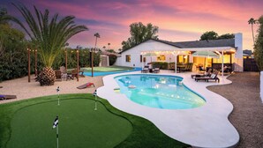 Un jardin avec une piscine, un terrain d’entraînement au golf et une maison dotée d’un espace salon extérieur.