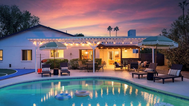 Un espace piscine avec des chaises longues et des parasols, une terrasse couverte et une maison à l’extérieur blanc.