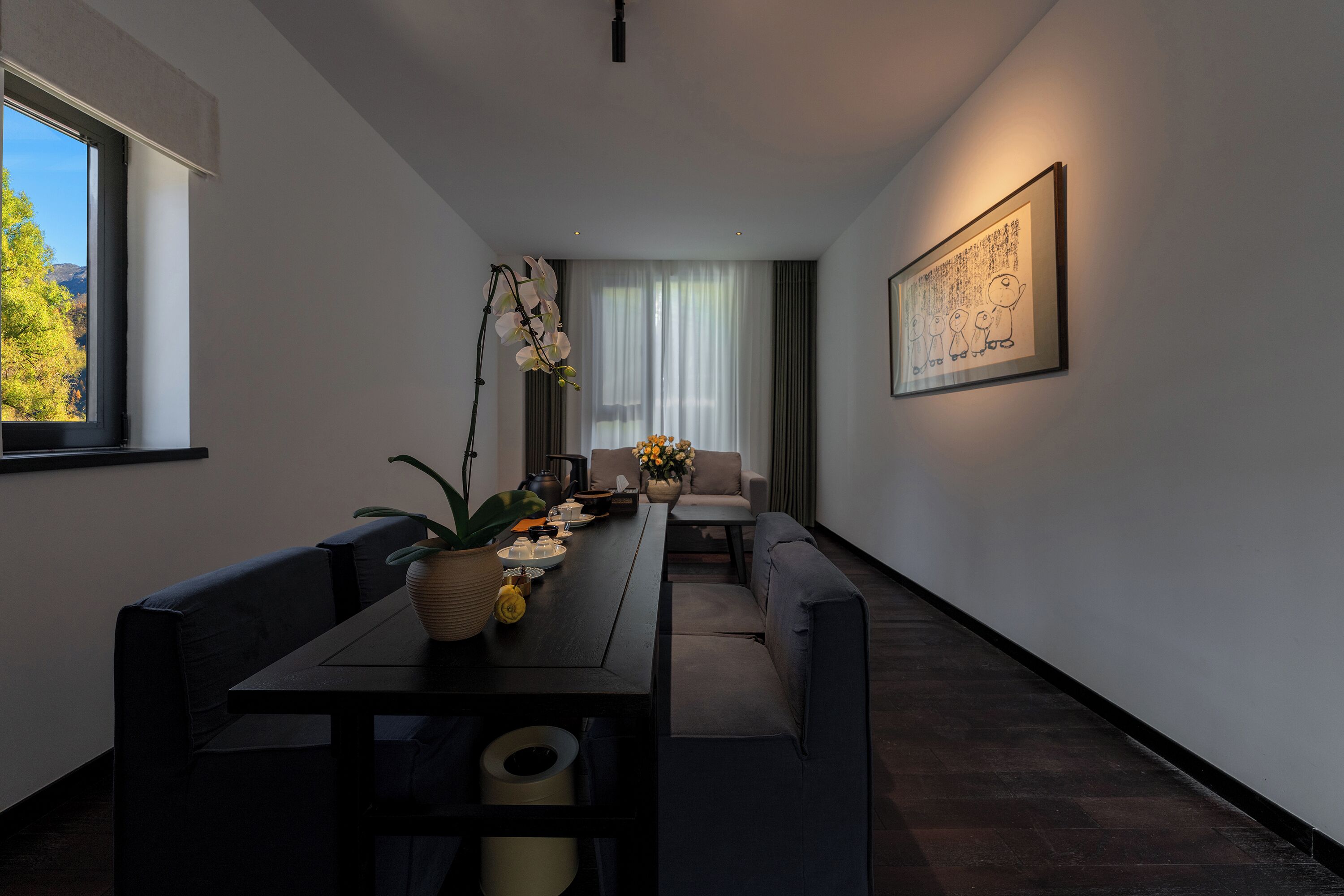A modern living room with a dining table, sofas, a potted plant, and a framed artwork on the wall.