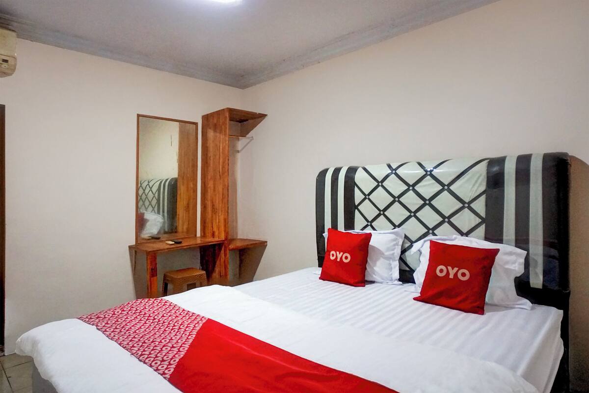 Ein Schlafzimmer mit einem Bett, zwei rote Kissen mit dem Aufdruck "OYO", einem hölzernen Nachttisch, einem Spiegel und einem Fenster mit Jalousien.