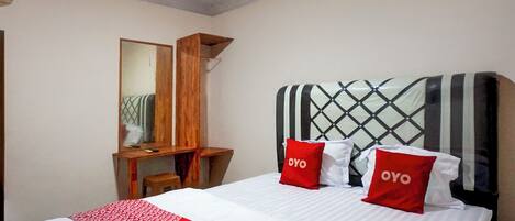 A bedroom with a bed, two red pillows with "OYO" text, a wooden nightstand, a mirror, and a window with blinds.