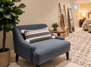 A room with a blue armchair, a potted plant, a small table with a plant and a candle, a mirror, and a bed with a patterned rug.