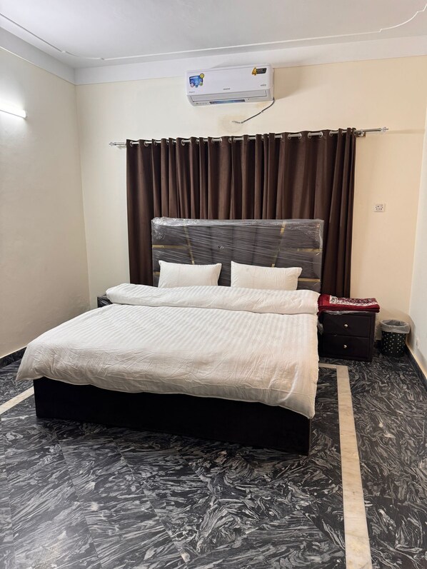 A neatly made bed with a headboard, a bedside table with a lamp, and a wall-mounted air conditioner. - Taj Villa (Islamabad)