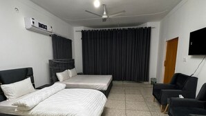 A hotel room with two beds, a ceiling fan, a TV, and a seating area with chairs. - Taj Villa (Islamabad)