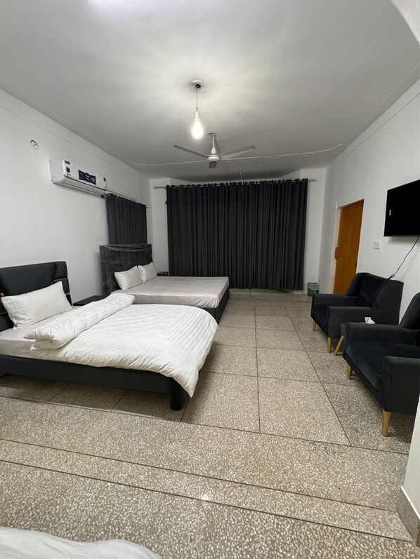 Habitación de hotel con dos camas, ventilador de techo, televisor y zona de estar con sillas.