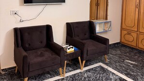 A room with two dark armchairs, a flat-screen TV mounted on the wall, a small side table with a box and a glass, and a wooden wardrobe with a mirror. - Taj Villa (Islamabad)