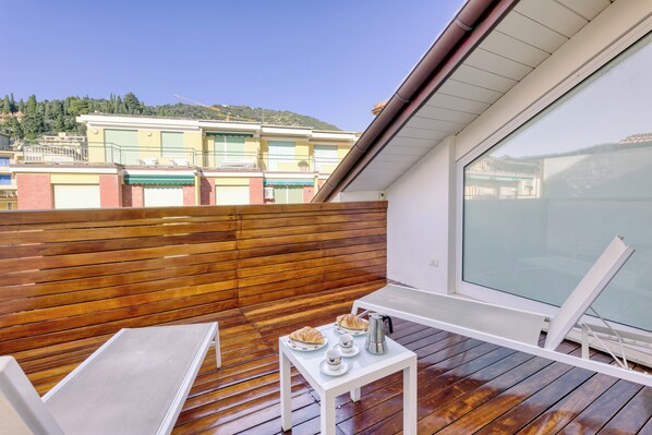 Un balcón con piso de madera, una mesa blanca con platos y tazas, y vistas a edificios al fondo.