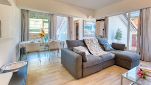 Ein modernes Wohnzimmer mit einem grauen Ecksofa, einem Glastisch und einem Essbereich mit weißem Tisch und Stühlen.