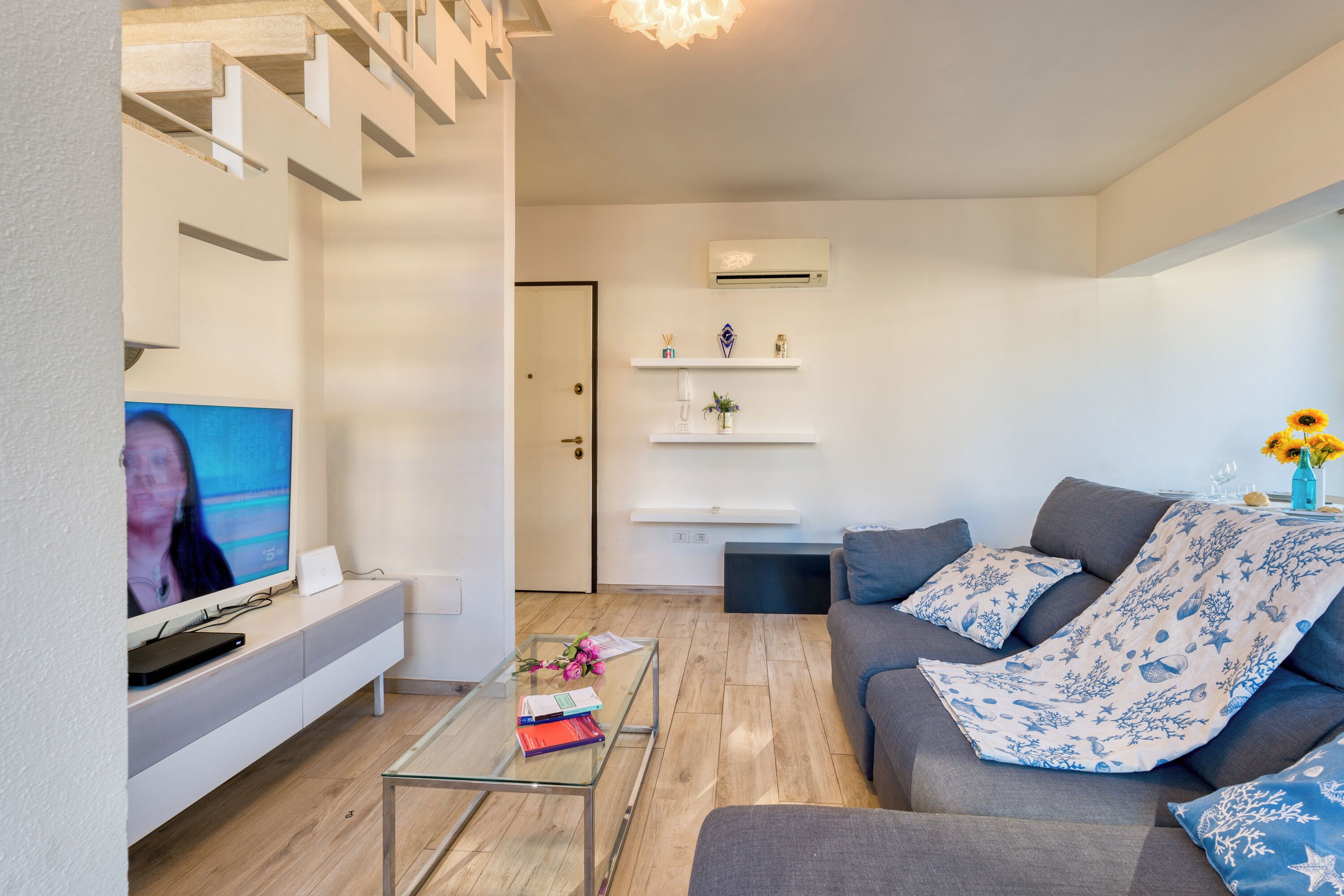 Un salon moderne avec un téléviseur à écran plat, une table basse en verre, un canapé gris et un escalier.