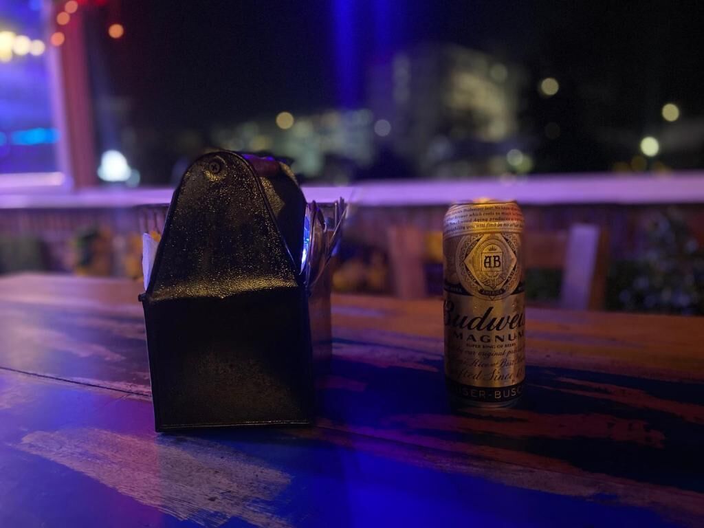 Una botella oscura de cerveza Budweiser Magnum y un objeto negro sobre una mesa, con un paisaje urbano borroso al fondo.