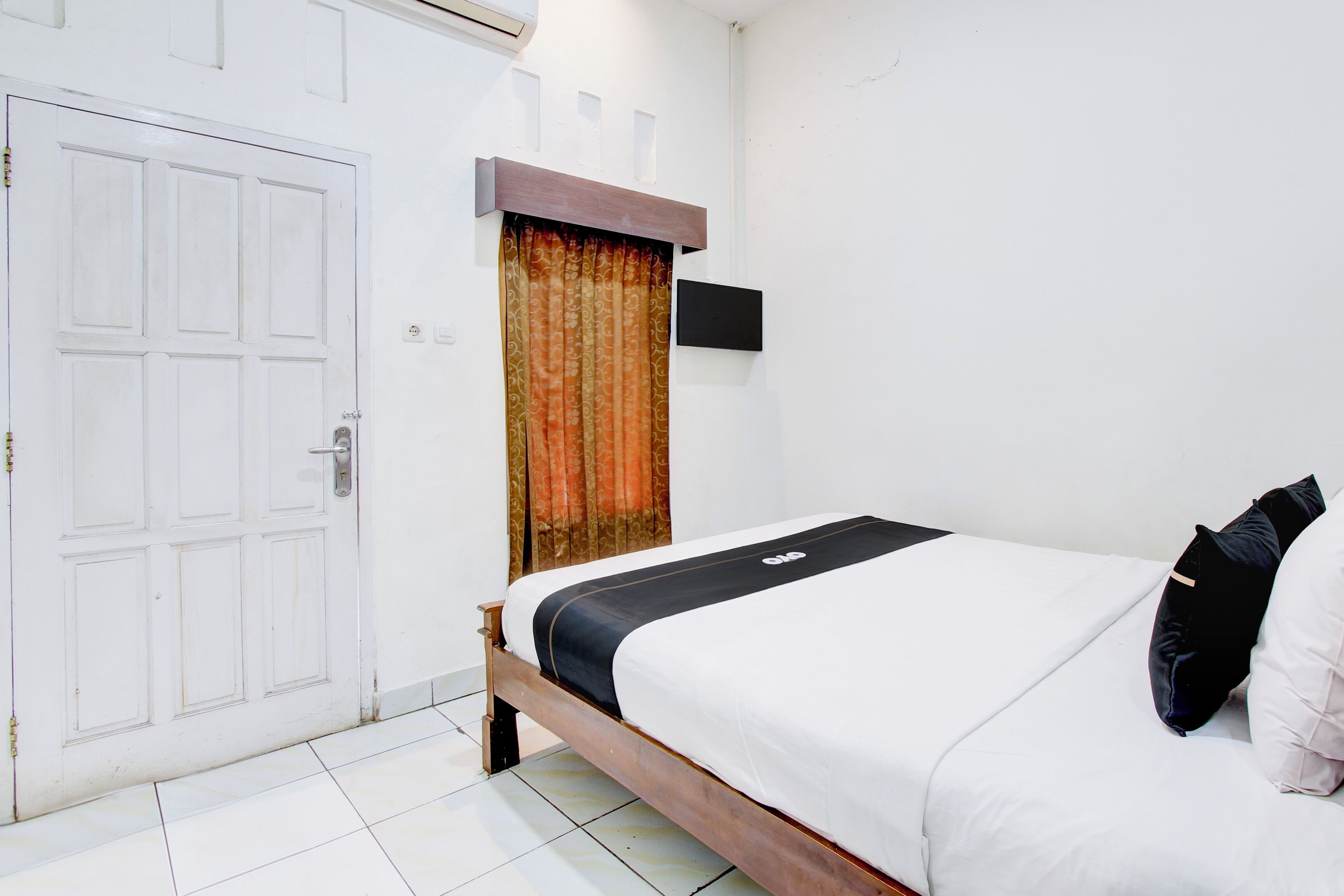 Un dormitorio con cama de madera, ropa de cama blanca, una cortina, un televisor montado y una puerta blanca.