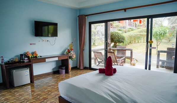 A hotel room with a bed, a desk with a TV, a small fridge, a trash bin, and a view of the outdoor area. - Chomview Lanta Water Park Resort (Ko Lanta)