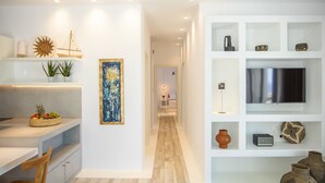 A modern kitchen with a wooden dining table, a built-in bench, and a fruit basket. There is a hallway leading to a bedroom with a bed and a nightstand. - Nice Villa at Kalafatis Beach (Mykonos)