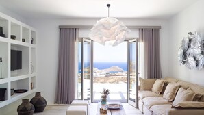 A modern living room with a large window, a sofa, a coffee table, and decorative items. - Nice Villa at Kalafatis Beach (Mykonos)