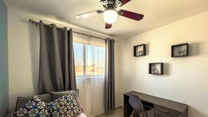 Quarto com cama, escrivaninha e cadeira. Há uma janela com cortinas, um ventilador de teto e quadros pendurados na parede.